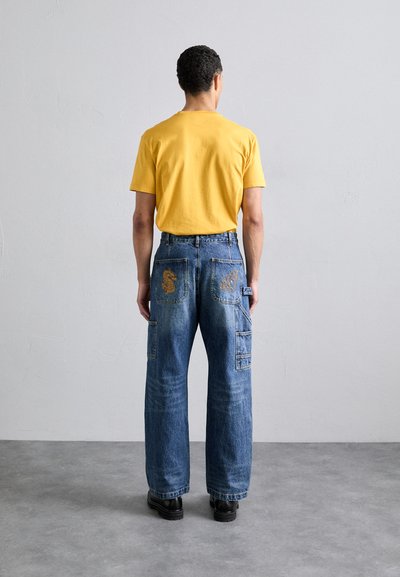 T-shirt en coton jaune associé à un jean en denim bleu baggy avec des motifs brodés sur les poches arrière. Des chaussures noires complètent le look.