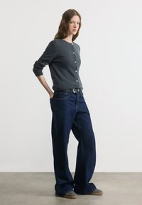 Γυναίκα στέκεται με τα χέρια στις τσέπες φορώντας σκούρο γκρι κουμπωτό ζακέτα, φαρδιά σκούρα μπλε τζιν, μαύρη ζώνη και γκρι αθλητικά παπούτσια σε γκρι πάτωμα.