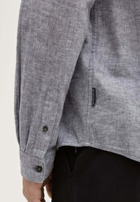 Chemise en lin grise à manches longues avec des poignets boutonnés, présentant un ourlet droit et une étiquette sur le côté, mettant en avant un tissu texturé.