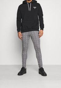 Felpa nera con cappuccio e logo bianco; leggings grigi a quadri; sneakers nere. Presenta una tasca frontale e stringhe sul cappuccio.