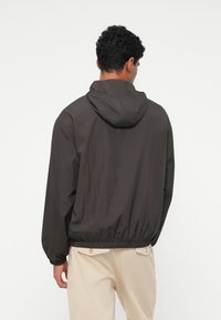 Giacca leggera con cappuccio di colore grigio scuro, dotata di polsini e vita elasticizzati. Texture liscia con dettagli minimi.