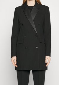 Kvinna iklädd en dubbelknäppt svart blazer med satinkragar och matchande svarta byxor, stående mot en enkel bakgrund.