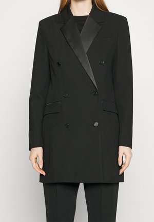 Frau in einem doppelt geknöpften schwarzen Blazer mit satinierten Revers und passenden schwarzen Hosen, stehend vor einem einfarbigen Hintergrund.