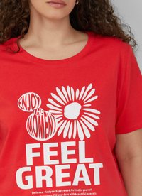 Czerwona bawełniana koszulka z białym napisem i kwiatowym wzorem, z wyraźnie nadrukowanymi hasłami "CIESZ SIĘ CHWILĄ" oraz "CZUJ SIĘ ŚWIETNIE".