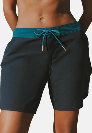 Mittelteil einer Person, die schwarze Badeshorts mit einem türkisfarbenen Bund und gebundenem Kordelzug trägt, stehend mit den Händen hinter dem Rücken.