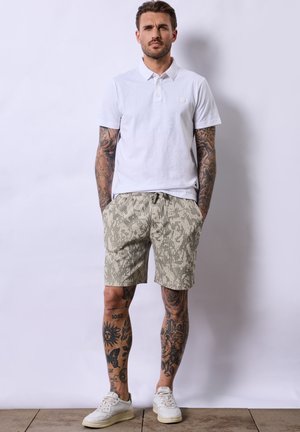Bílá polo košile s krátkým rukávem a decentním logem, kombinovaná se světle šedými šortkami se vzorem camouflage. Celkový vzhled doplňují bílé tenisky.