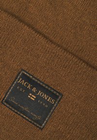 Tissu de pull en laine brun tricoté avec une finition texturée, arborant une étiquette foncée avec du texte doré et des coutures, affichant le nom de la marque et la date de création.