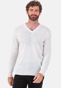 Leichter, weißer Pullover mit V-Ausschnitt aus weichem, strukturiertem Material, mit langen Ärmeln und gerippten Bündchen. Über dunkler Hose getragen.