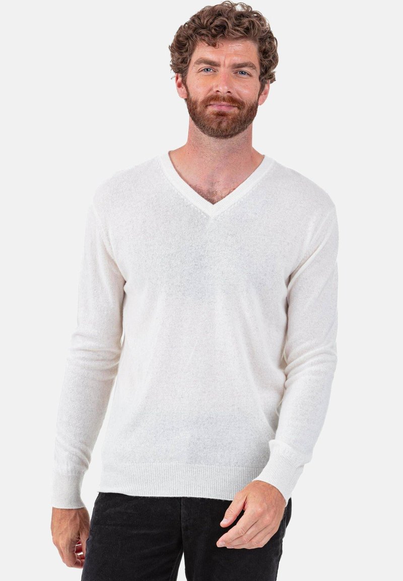 Leichter, weißer Pullover mit V-Ausschnitt aus weichem, strukturiertem Material, mit langen Ärmeln und gerippten Bündchen. Über dunkler Hose getragen.