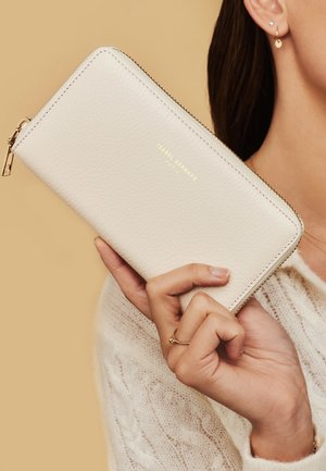 Witte leren portemonnee met een textuur, gouden rits en het merklogo in goud gedrukt. Slanke rechthoekige vorm vastgehouden in de hand.
