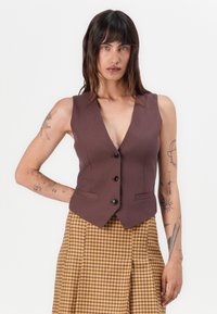 Gilet marron ajusté avec un décolleté en V profond, cinq boutons et deux poches latérales, associé à une jupe à carreaux jaunes et bruns.