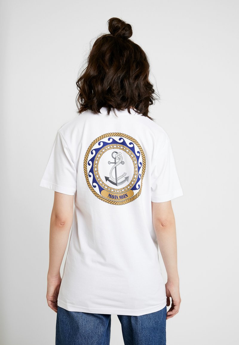 Hvid t-shirt med et cirkulært grafisk design på bagsiden, der har et ankeromrids omgivet af gyldent reb og blå bølger, med teksten "MOIN MOIN."