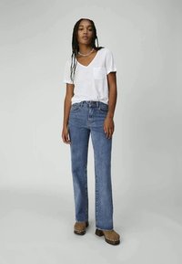 Woman with long braided hair wearing a white V-neck T-shirt, blue straight-leg jeans, brown clogs, and a silver chain necklace.