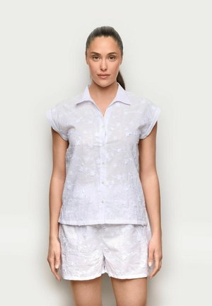 Donna con capelli castani raccolti in una coda di cavallo, che indossa una camicia bianca a maniche corte con ricami floreali e pantaloncini abbinati, in piedi davanti a uno sfondo neutro.