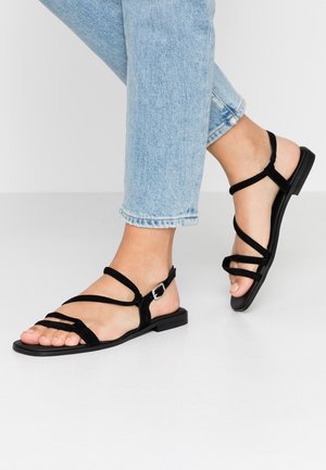 Sorte ruskind flade sandaler med flere slanke stropper. Har et justerbart ankelsluk og en simpel flad sål. Båret med lyseblå jeans.