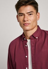 Bordeaux knoopshirt met een klassieke kraag, witte knopen en een subtiele logodetail op de borst, gelaagd over een wit shirt.