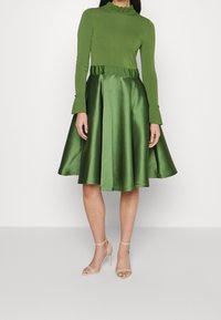 Mujer llevando un top de punto de manga larga verde y una falda midi acampanada verde brillante con sandalias de tacón alto beige, de pie sobre un fondo claro.