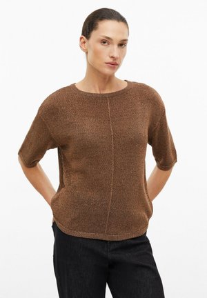 Frau trägt einen braunen kurzärmeligen Strickpullover mit Mittelnaht, kombiniert mit einer schwarzen Hose, steht vor einem weißen Hintergrund.
