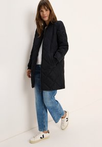 Zwarte gewatteerde jas met ritsluiting, zijzakken en elastische manchetten, gecombineerd met lichtblauwe wide-leg jeans en witte sneakers.