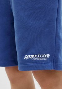 Blå bomullsshorts med en mjuk textur, med en vit "project core"-logotyp vid fållen och en enkel, rak design.