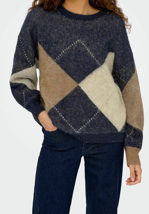 Femme portant un pull duveteux multicolore avec des motifs losanges bleu marine, beige et crème et un jean bleu foncé.