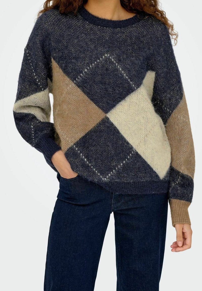 Femme portant un pull duveteux multicolore avec des motifs losanges bleu marine, beige et crème et un jean bleu foncé.