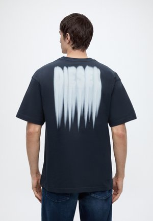 Homme aux cheveux courts et foncés portant un t-shirt noir avec "DIESEL" en grandes lettres blanches délavées dans le dos, associé à un jean foncé.