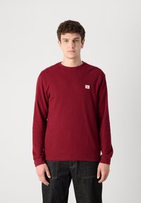 Bordeaux langemouwen shirt met ribbeltextuur, voorzien van een klein logo op de borst. Gecombineerd met zichtbare donkere spijkerbroek.