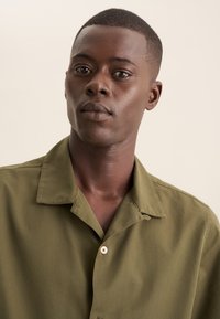 Camicia a maniche corte verde oliva in cotone. Presenta un colletto classico e un singolo bottone chiaro, con una texture liscia.