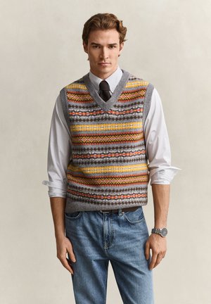 Jeune homme aux cheveux bruns coiffés, portant un gilet en tricot à motifs sur une chemise blanche et une cravate noire, associé à un jean bleu et une montre-bracelet.
