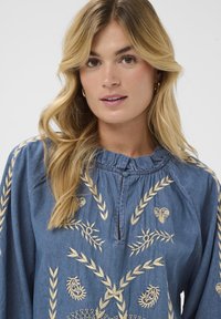 Blondide naine, kellel on seljas sinine särk, millel on beeži värvi tikandid lehtede ja paisley mustritega ning volangidega krae, valge tausta ees.