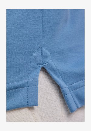 Primo piano di tessuto azzurro chiaro che mostra una cucitura rinforzata e una piccola spacco laterale con cuciture dettagliate sul bordo di un indumento.