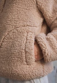 Lyserød fleece-hættetrøje med blød, behåret overflade og en lomme foran. Manchetter og kant er foret med et blødt, plyssede materiale.