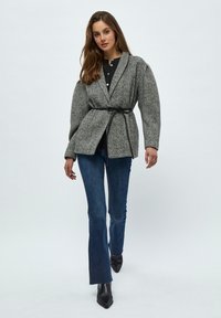 Grå hængekøje blazer med bælte i taljen, suppleret af en sort top og blå flagrende jeans; modellen har spidse sorte højhælede sko på.