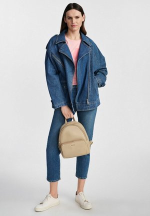 Kobieta w dużej niebieskiej kurtce dżinsowej i dżinsach, trzymająca beżowy plecak, ubrane w białe trampki i różowy top, stojąca na tle jednolitego tła.