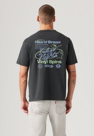 Mann mit kurzen Haaren trägt ein schwarzes T-Shirt mit Grafik, auf dem "Haus of Strauss", eine Vinyl-Schallplatte und der Schriftzug "Indigo Nights" zu sehen sind, kombiniert mit hellen Jeans.