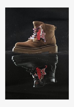 Stivale in suede marrone con lacci colorati e portachiavi rosso "Stranger Things" appeso di lato, riflesso su una superficie nera lucida.