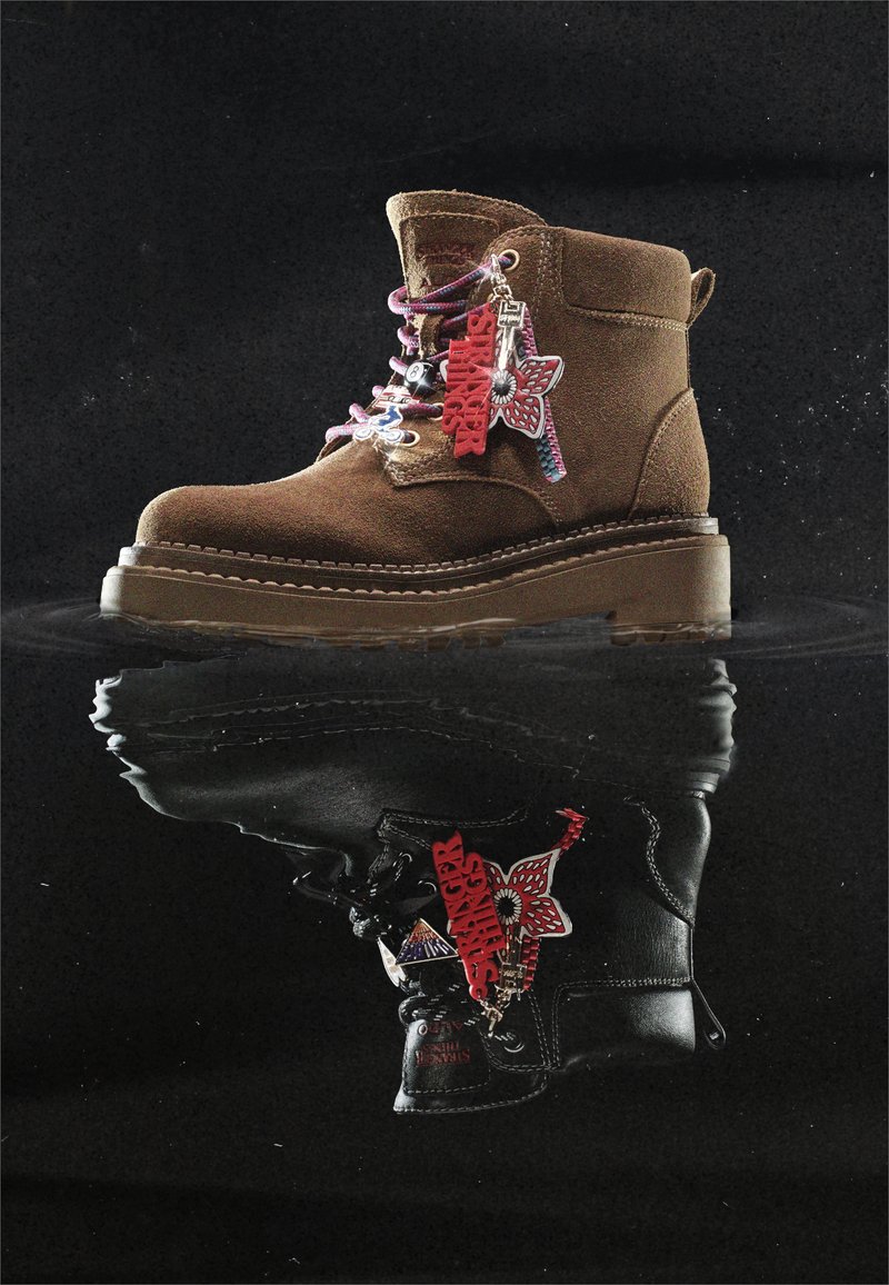 Stivale in suede marrone con lacci colorati e portachiavi rosso "Stranger Things" appeso di lato, riflesso su una superficie nera lucida.
