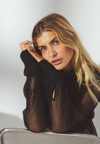 Sort plisseret gennemsigtig bluse med svulmende ærmer, der har knapdetaljer og en glat tekstur, båret af en model med bølget blond hår.