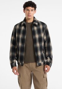 Crna i bež karirana jakna s patentnim zatvaračem, ovratnik dizajna i džepom s jedne strane, nosi se preko maslinasto zelene košulje.