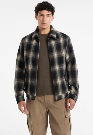 Czarna i beżowa kurtka w kratę z zamkiem, kołnierzowym fasonem i kieszenią po jednej stronie, noszona na oliwkowej koszuli.