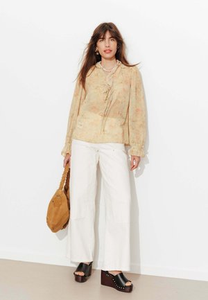 Femme aux longs cheveux bruns portant un chemisier beige à fleurs, un pantalon blanc à jambes larges, des sandales compensées noires, et tenant un sac à main tressé couleur cuir clair.