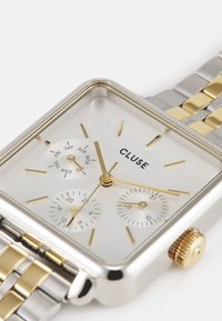 Montre en métal argenté carrée avec des accents dorés, cadran affichant le jour et la date, bracelet en maillons argentés et dorés, éléments de design minimalistes.