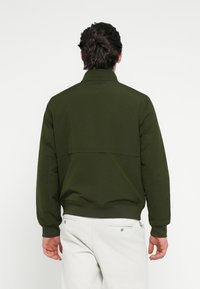 Jachetă bomber verde măsliniu cu guler în picioare, material texturat, mansete și tiv striate. Detalii remarcabile ale cusăturilor pe partea din spate.