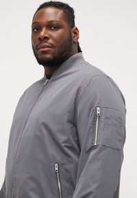 Chaqueta bomber gris con cremallera frontal, cuello acanalado y múltiples bolsillos con cremallera en las mangas y laterales. Textura suave.