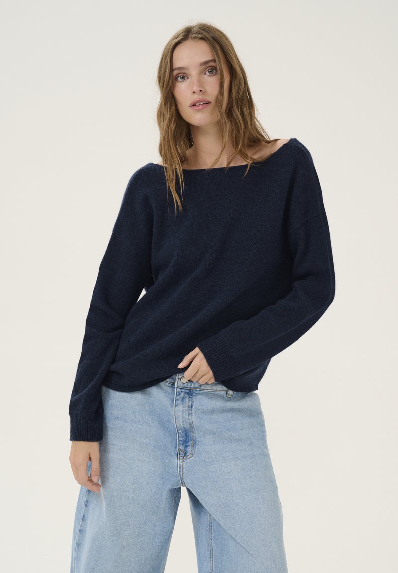 Tengerészkék, oversized kötött pulóver széles nyakkivágással és hosszú ujjakkal, világoskék farmernadrággal, amely egyedi szabással rendelkezik.