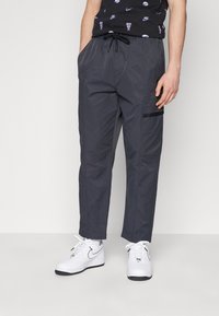 Grijze cargobroek met elastische tailleband, verstelbaar trekkoord, zijzakken en zwarte accenten. Draagt met witte sneakers.