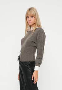 Blonde vrouw in een grijze gebreide trui over een wit shirt en zwarte leren broek, die zijwaarts poseert tegen een witte achtergrond.
