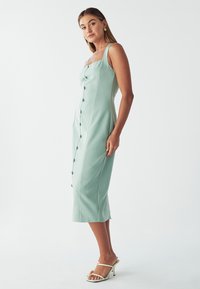 Mintgrøn midi-kjole med tætsiddende liv, firkantet halsudskæring og knapdetaljer. Har tynde stropper og en glat, elegant tekstur.