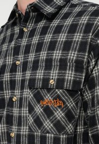 Μαύρο και άσπρο καρό φανελένιο πουκάμισο με μπεζ κουμπιά και τσέπη στο στήθος με πορτοκαλί κεντημένη λεπτομέρεια. Απαλή υφή, κανονικός γιακάς.